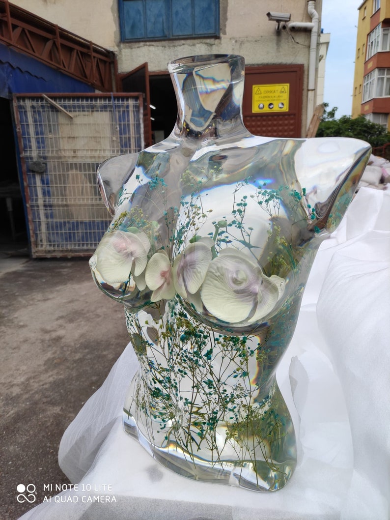 Puede incluir: Escultura de busto de resina transparente con flores de orqu&iacute;deas blancas y delicado follaje verde y azul. El busto es una forma de torso. La escultura se exhibe sobre una superficie blanca.