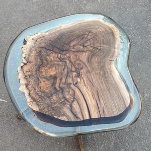 May include: A unique, handcrafted coffee table with a live edge wood slab top. The wood is a rich brown color with intricate grain patterns, encased in a clear resin. The table is supported by brass-colored metal legs.