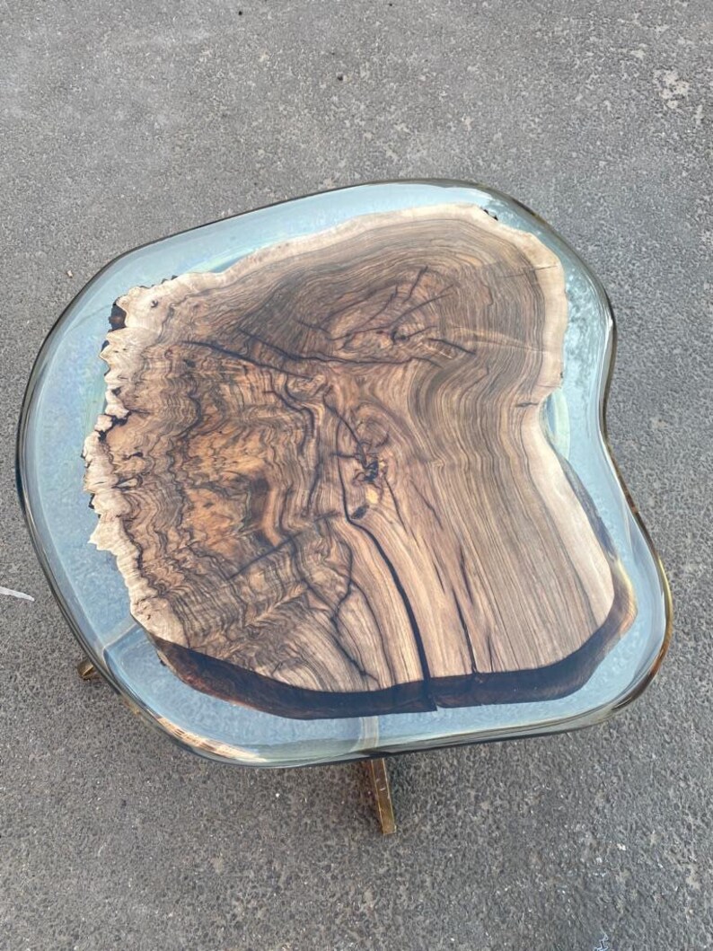 May include: A unique, handcrafted coffee table with a live edge wood slab top. The wood is a rich brown color with intricate grain patterns, encased in a clear resin. The table is supported by brass-colored metal legs.