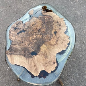 May include: A unique, handcrafted coffee table with a live edge wood slab encased in clear resin. The wood features rich brown tones and intricate grain patterns. The table is supported by brass-colored legs.