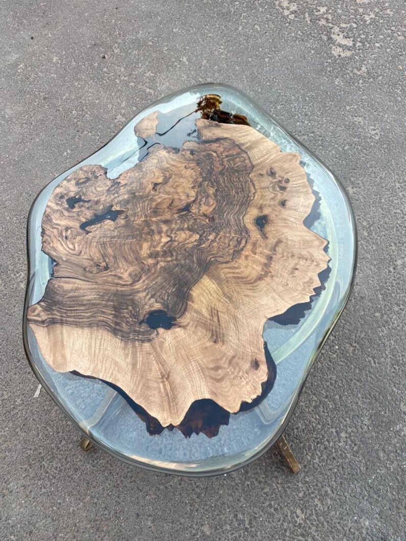 May include: A unique, handcrafted coffee table with a live edge wood slab encased in clear resin. The wood features rich brown tones and intricate grain patterns. The table is supported by brass-colored legs.