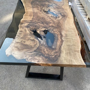 May include: A large, rectangular wooden table with a live edge and a black metal base. The tabletop features a natural wood grain pattern with areas of dark blue resin. The table measures approximately 96 inches long and 40 inches wide.