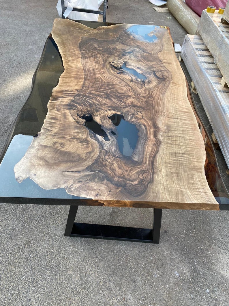 May include: A large, rectangular wooden table with a live edge and a black metal base. The tabletop features a natural wood grain pattern with areas of dark blue resin. The table measures approximately 96 inches long and 40 inches wide.
