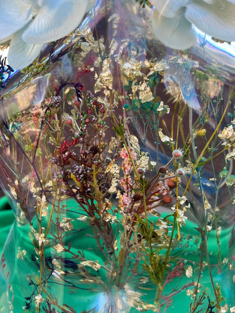 Puede incluir: Una escultura de resina transparente con flores secas e inclusiones de pan de oro. La escultura tiene forma de busto, sobre un fondo verde. Las flores son de varios colores, incluyendo rosa, marr&oacute;n y blanco.