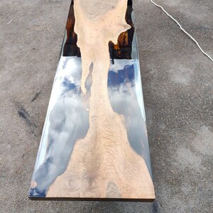 May include: A rectangular wooden table with a clear epoxy resin river. The wood has a light brown colour with a unique grain pattern. The resin reflects the sky, creating a cloudy effect. The table is approximately 152 cm long.