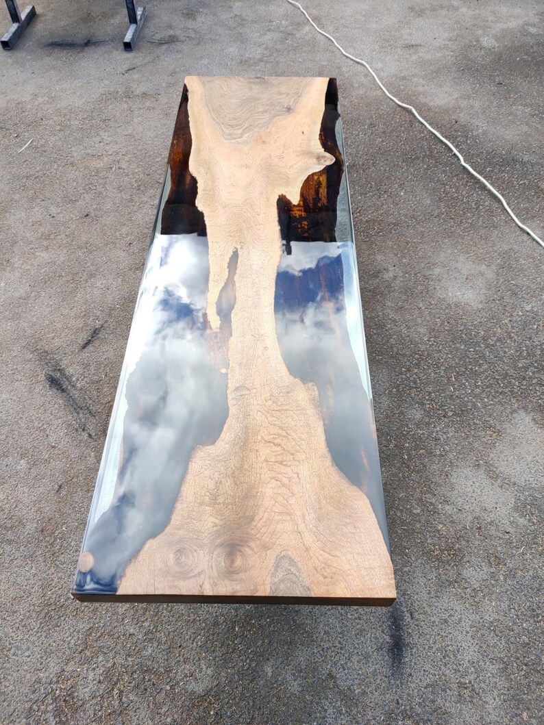 May include: A rectangular wooden table with a clear epoxy resin river. The wood has a light brown colour with a unique grain pattern. The resin reflects the sky, creating a cloudy effect. The table is approximately 152 cm long.