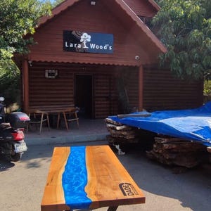 May include: A wooden table with a blue resin river design. The table has a dark metal base and is displayed outside a shop with the sign "Lara Wood's". The table is approximately 60 inches long.
