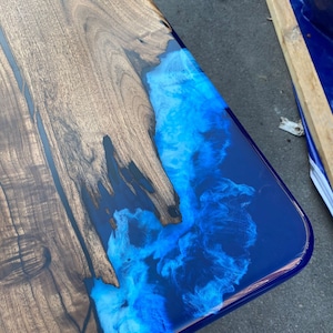 Könnte beinhalten: Handgefertigter Holztisch mit auffälligem Epoxidharz-Design. Der Tisch hat auf einer Seite eine dunkle Holzmaserung, die durch einen lebendigen blau-weißen Harzfluss kontrastiert wird. Das Harz hat ein wirbelndes, wolkenartiges Aussehen. Die Tischlänge beträgt ca. 100-120 cm.