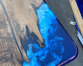 May include: A handcrafted wooden table with a striking epoxy resin design. The table features a rich, dark wood grain on one side, contrasted by a vibrant blue and white resin river flowing across the corner. The resin has a swirling, cloud-like appearance.