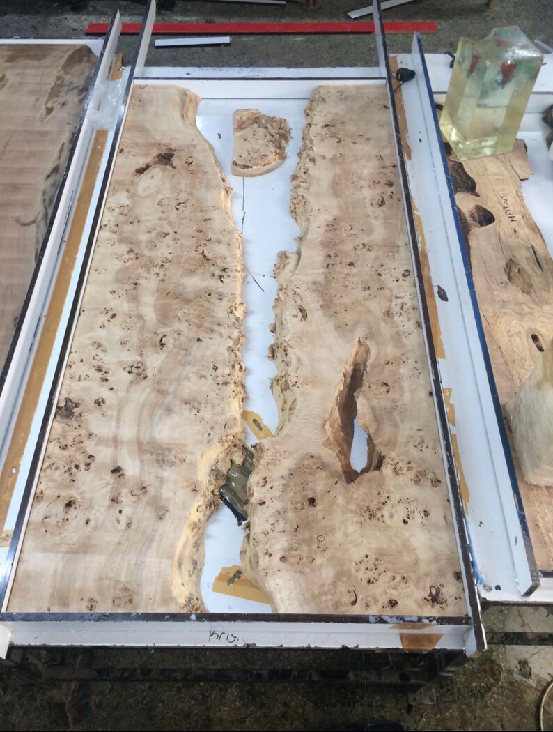 Pode incluir: Uma grande laje de madeira inacabada com uma superf&iacute;cie castanha clara e salpicada. A madeira tem uma borda natural e irregular e um canal central. Est&aacute; colocada dentro de uma moldura branca e met&aacute;lica, sugerindo uma mesa ou bancada em andamento.