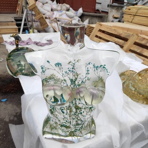 Puede incluir: Una escultura de busto de resina transparente con inclusiones bot&aacute;nicas. La escultura presenta un cuello rectangular y un torso con elementos florales y vegetales. El busto se exhibe sobre una superficie blanca. El busto mide unos 30 cm de altura.
