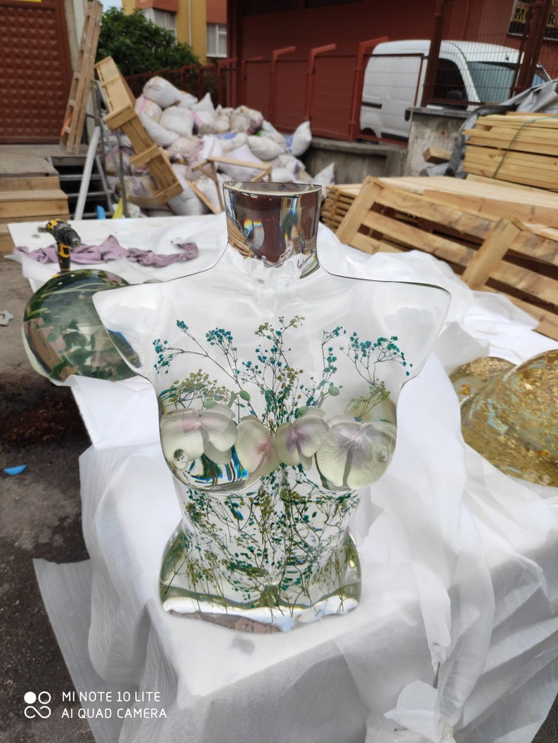 Puede incluir: Una escultura de busto de resina transparente con inclusiones bot&aacute;nicas. La escultura presenta un cuello rectangular y un torso con elementos florales y vegetales. El busto se exhibe sobre una superficie blanca. El busto mide unos 30 cm de altura.