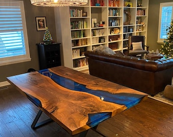 May include: A handcrafted wooden dining table with a blue resin river design. The table features a dark wood grain and a glossy blue center, supported by black metal legs. The table is in a well-lit living room.