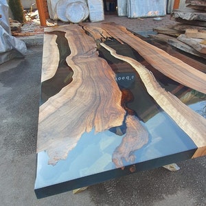 May include: A large, rectangular wooden table with a clear epoxy resin river. The table features natural wood grain patterns and a dark border. The table is approximately 96 inches long.