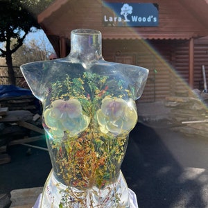 Puede incluir: Una escultura de torso de resina transparente con flores secas, hojas y una concha incrustadas. La escultura presenta dos grandes acentos florales de color claro y detalles de pan de oro. La escultura se exhibe sobre una superficie blanca.