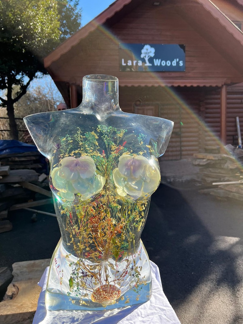 Puede incluir: Una escultura de torso de resina transparente con flores secas, hojas y una concha incrustadas. La escultura presenta dos grandes acentos florales de color claro y detalles de pan de oro. La escultura se exhibe sobre una superficie blanca.