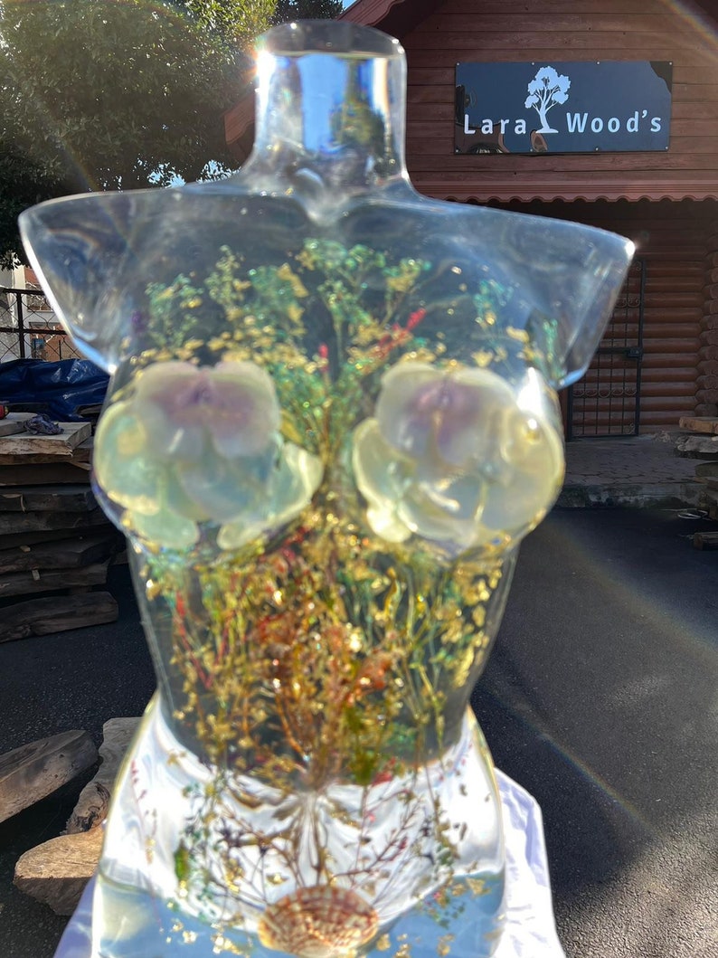 Puede incluir: Una escultura de resina transparente con forma de torso, llena de flores secas y hojuelas doradas. La escultura presenta dos formas de flores claras y una concha en la base. El fondo muestra un edificio de madera con el texto "Lara Wood's".