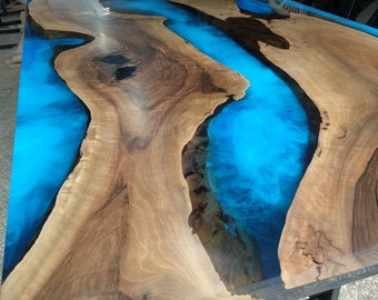 May include: A large wooden table with a river of bright blue resin running through the center. The wood has a natural grain pattern with varying shades of brown. The table is supported by a dark metal base.