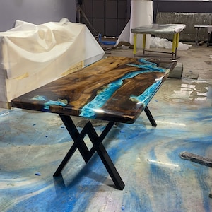 Könnte beinhalten: Ein rechteckiger Holztisch mit glänzendem Finish. Die Tischplatte zeigt einen Fluss aus blauem Harz, der durch die Mitte des Holzes fließt. Der Tisch hat schwarze Metallbeine in X-Form.