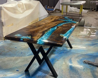 May include: A rectangular wooden table with a glossy finish. The tabletop features a river of blue resin flowing through the center of the wood. The table has black metal legs in an X-shape.