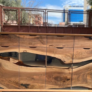 May include: A wooden cabinet with a natural wood grain finish. The cabinet has multiple drawers and doors with a dark, glossy inlay. The design features a modern aesthetic with a focus on natural materials.
