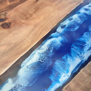 Könnte beinhalten: Nahaufnahme eines Holztisches mit einem Fluss aus blauem Harz. Das Holz hat eine natürliche Maserung und eine warme braune Farbe. Das Harz ist tiefblau mit weißen Wirbeln, die an Meereswellen erinnern.