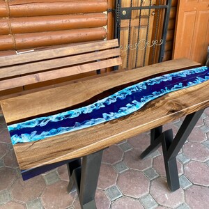 May include: A wooden river table with a dark wood grain finish and a blue epoxy resin river. The table has black metal legs. The river is a vibrant blue with white accents, creating a flowing water effect.