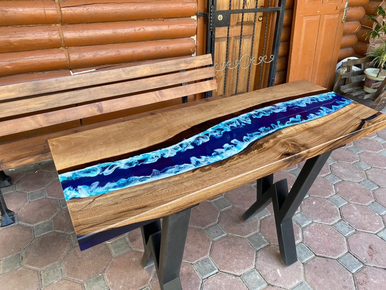 May include: A wooden river table with a dark wood grain finish and a blue epoxy resin river. The table has black metal legs. The river is a vibrant blue with white accents, creating a flowing water effect.