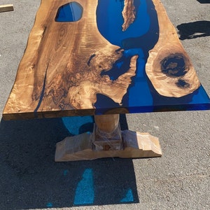 May include: A handcrafted wooden table with a live edge and a blue epoxy resin river. The table has a natural wood grain finish and sits on a pedestal base. The table is approximately 72 inches long.