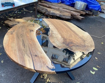 May include: A round wooden table with a black epoxy resin center. The table features a natural wood grain pattern and is supported by black metal legs. The table's diameter is approximately 48 inches.