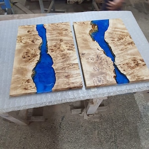 May include: Two wooden slabs with a river of blue resin running through them. The wood has a light brown color with a unique grain pattern. The blue resin creates a striking contrast, resembling a flowing river. The slabs are likely for a table or decorative piece.