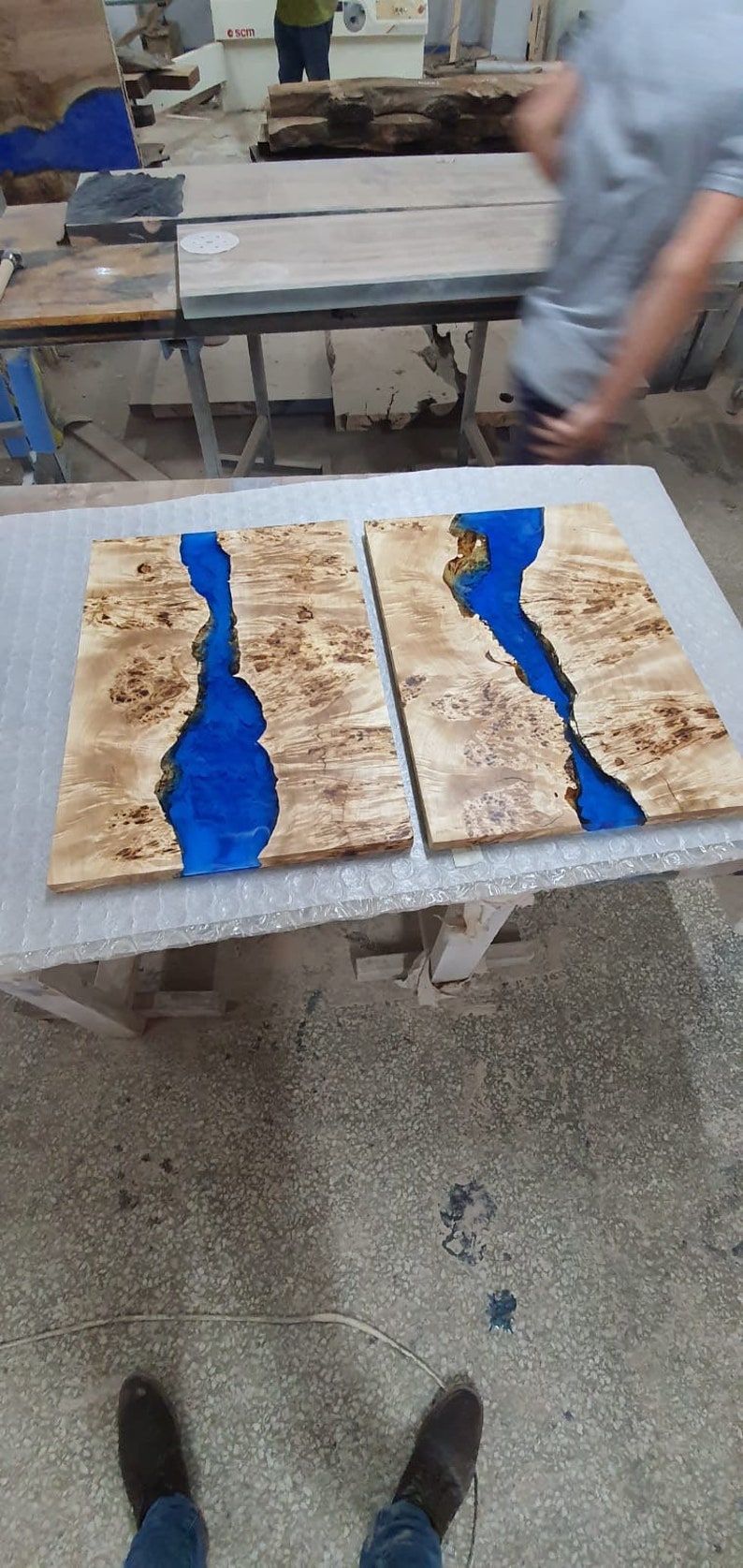 May include: Two wooden slabs with a river of blue resin running through them. The wood has a light brown color with a unique grain pattern. The blue resin creates a striking contrast, resembling a flowing river. The slabs are likely for a table or decorative piece.