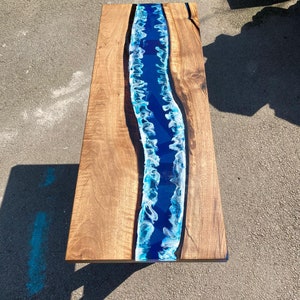 May include: A rectangular wooden coffee table with a river of blue resin running down the center. The wood has a natural grain and a warm brown color. The resin river has white and blue accents, creating a flowing water effect.