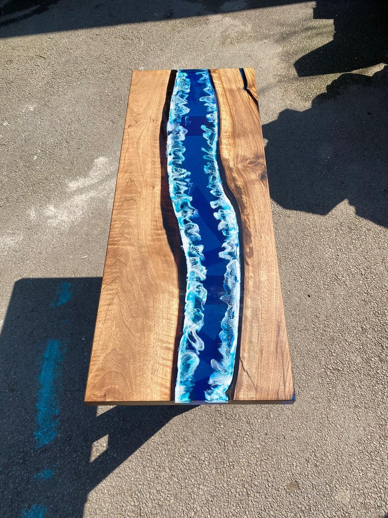 May include: A rectangular wooden coffee table with a river of blue resin running down the center. The wood has a natural grain and a warm brown color. The resin river has white and blue accents, creating a flowing water effect.