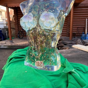Puede incluir: Una escultura de torso de resina transparente con flores incrustadas, una concha marina y copos de oro. La escultura presenta dos acentos florales de color p&uacute;rpura claro y blanco y se exhibe sobre una tela de vell&oacute;n verde.
