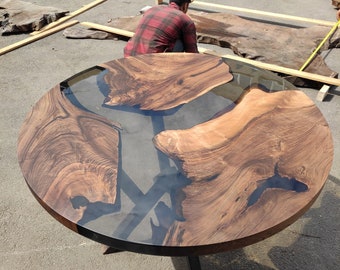 May include: A round wooden coffee table with a dark brown finish and a clear epoxy resin center. The table features a unique design with natural wood grain patterns and black metal legs. The table is approximately 48 inches in diameter.