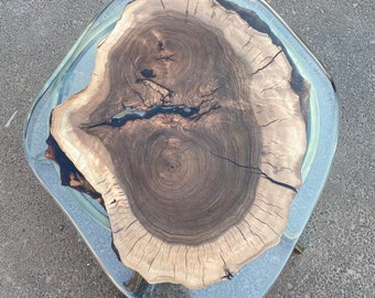 May include: A unique, handcrafted coffee table with a live edge wood slab encased in clear resin. The wood features a natural grain pattern with shades of brown and tan. The table has a clear, rounded edge and sits on gold-colored legs.
