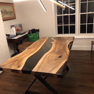 Könnte beinhalten: Ein großer, rechteckiger Holztisch mit einem dunklen, glänzenden Epoxidharzfluss in der Mitte. Der Tisch hat eine natürliche Holzmaserung und steht auf einem schwarzen Metallgestell. Der Tisch ist etwa 244 cm lang.