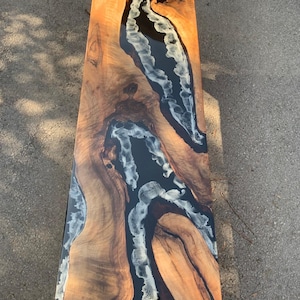 May include: A rectangular wooden table with a dark brown epoxy river design. The wood has a natural grain with shades of light and dark brown. The river is black with white and gray accents, creating a unique, artistic look.