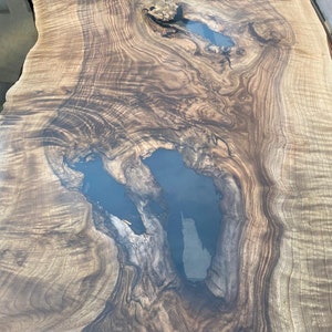 May include: A close-up view of a large, live edge wooden table. The wood grain is a mix of light and dark brown, with areas of dark blue resin filling natural voids. The surface is smooth and polished, highlighting the natural patterns.
