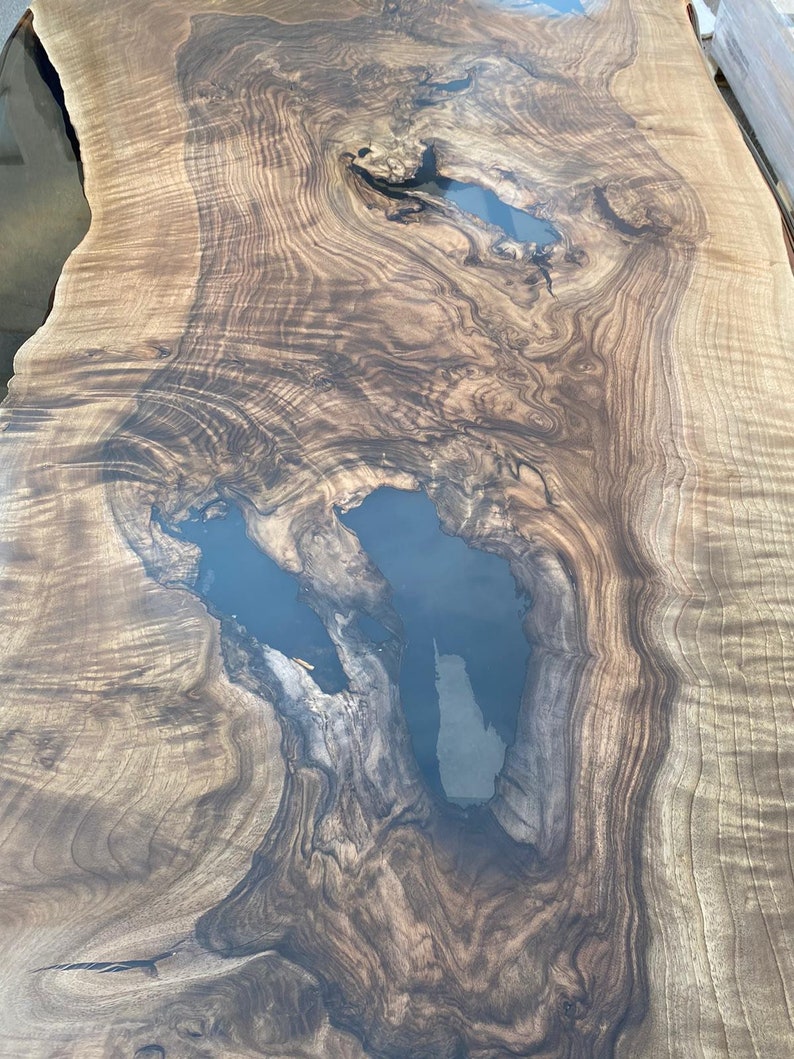 May include: A close-up view of a large, live edge wooden table. The wood grain is a mix of light and dark brown, with areas of dark blue resin filling natural voids. The surface is smooth and polished, highlighting the natural patterns.