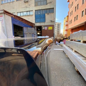 May include: A low-angle shot of a black, glossy car bumper reflecting the surrounding buildings and sky. The bumper curves slightly, with a smooth, polished surface. The background includes industrial buildings and a sunny sky.