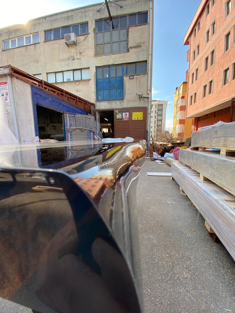 May include: A low-angle shot of a black, glossy car bumper reflecting the surrounding buildings and sky. The bumper curves slightly, with a smooth, polished surface. The background includes industrial buildings and a sunny sky.