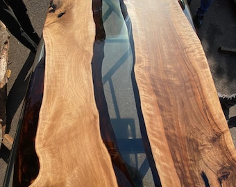 May include: A large, rectangular wooden table with a clear epoxy river running through the center. The table features two live edge wooden slabs with a natural wood grain pattern. The wood is a light brown color with darker brown accents.