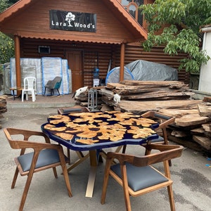 May include: A wooden table with a dark blue epoxy resin top featuring a natural wood grain pattern, surrounded by four wooden chairs with gray seats. The table is supported by a gold-colored metal base. The business sign reads "Lara Wood's".