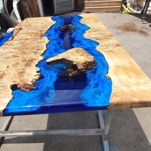 Op de afbeelding: Een grote, rechthoekige houten tafel met een rivier van blauwe epoxyhars in het midden. De tafel wordt ondersteund door een zilveren metalen frame. Het hout heeft een lichtbruine kleur.