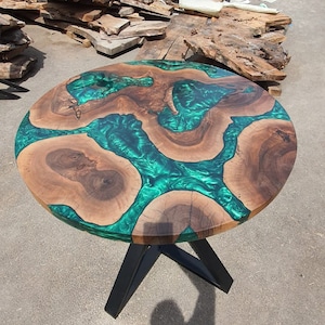 May include: Round wooden table with a unique design. The tabletop features a combination of natural wood and vibrant green epoxy resin. The table is supported by a black metal base, creating a modern aesthetic. The table measures approximately 40 inches in diameter.
