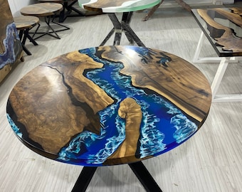 May include: A round wooden table with a river-like blue epoxy resin design. The table has a dark wood grain pattern and sits on black metal legs. Other tables with similar designs are in the background.