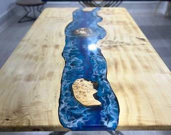 May include: A large wooden table with a river-like blue epoxy resin center. The table has a light wood grain finish and a unique design. The table is supported by a metal base.