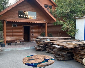 May include: A round wooden table with a blue and green epoxy resin design sits in front of a wooden building. The table has a metal base. The building has a sign that reads "Lara Wood's".